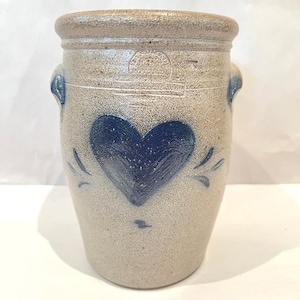 Puede incluir: Un tarro de gres vintage con un motivo de corazón azul. El tarro tiene un cuerpo gris claro, un borde gris más oscuro y asas. El corazón está colocado en el centro, con adornos decorativos. Un artículo de cocina clásico.