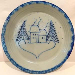 May include: A circular, off-white ceramic plate with a blue border, featuring a winter scene. The design includes a house, trees, and falling snow, all in blue. The plate has a rustic, handmade aesthetic.