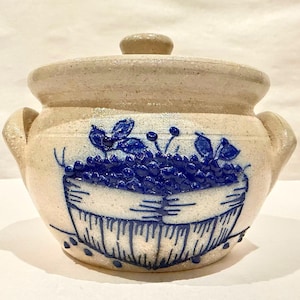 Puede incluir: Un cuenco de cerámica beige con tapa y dos asas. El cuenco presenta un diseño pintado en azul de una cesta llena de uvas. La tapa tiene un pequeño pomo para facilitar su extracción.