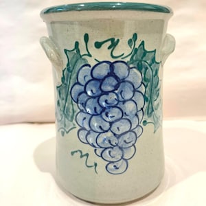 May include: A ceramic utensil holder with a light green base, adorned with a blue grape cluster and green leaves. The rim and handles are also green. This kitchenware item is ideal for storing cooking tools.