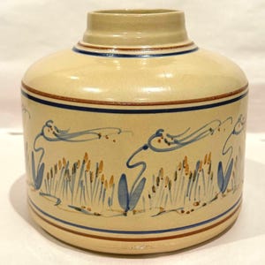 May include: A cylindrical ceramic vase with a cream-colored glaze, featuring a hand-painted blue and yellow floral motif around the center. The vase has a short, narrow neck and is accented with thin horizontal blue and brown bands at the top and base.