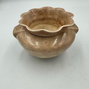 May include: A speckled, light brown ceramic bowl with a ruffled rim. The bowl has a rounded base and two small handles on either side. The interior is a lighter shade of brown.