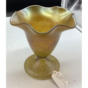 Puede incluir: Un jarrón de vidrio iridiscente antiguo con borde ondulado y base bulbosa. El jarrón tiene una superficie moteada y texturizada con un tono amarillo dorado. Una etiqueta adjunta a la base dice "Loetz Silberiris 1914."