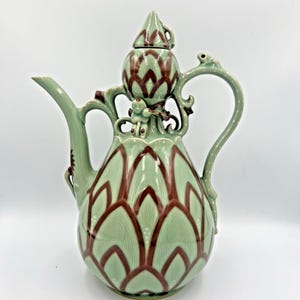 May include: A celadon-colored ceramic pitcher with a bulbous body and a long spout. The pitcher is decorated with a repeating pattern of maroon-colored floral designs. The handle is curved and features a small frog. The lid is cone-shaped and has a small handle.