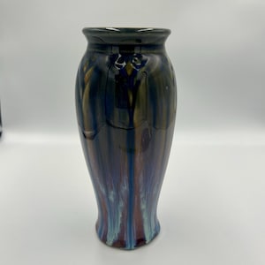 May include: A tall, ceramic vase with a flared base and a dark, glossy finish. The vase features a blend of colors, including blue, brown, and purple, with vertical streaks creating a flowing effect. The rim is dark, and the overall shape is elegant.