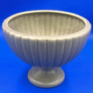 May include: A light grey, speckled ceramic bowl with a vertical ribbed texture on the exterior. The bowl features a wide, open top and is supported by a circular pedestal base, creating a classic, rounded silhouette for this piece.