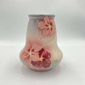 May include: A ceramic vase with a flared top and a bulbous body. The vase is painted with a gradient of colors, transitioning from a light blue at the top to a soft pink at the bottom. It features embossed pansy flowers in shades of pink and burgundy.