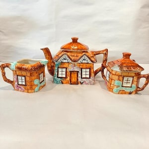 May include: A ceramic tea set consisting of a teapot, a creamer, and a sugar bowl, all shaped like small cottages with thatched roofs. The pieces feature brown brick patterns, white window frames, and painted green and purple floral accents.