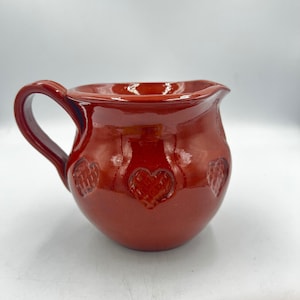 Puede incluir: Una jarra de cerámica roja brillante con asa y boquilla. La jarra presenta tres diseños de corazón en relieve alrededor del cuerpo. La cerámica hecha a mano es adecuada para servir bebidas.