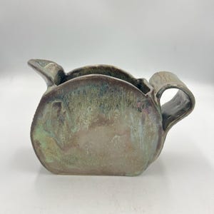Könnte beinhalten: Ein handgefertigter Keramikkrug mit einer einzigartigen, organischen Form. Der Krug hat einen Ausguss und einen gebogenen Griff. Die Glasur hat eine Mischung aus Erdtönen, darunter Grün, Braun und Hauch von Lila, wodurch ein strukturiertes, schillerndes Finish entsteht. Der Krug eignet sich zum Servieren von Getränken.
