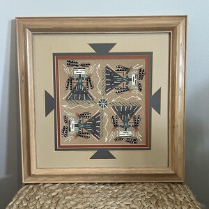 May include: Framed artwork featuring a geometric design with four quadrants. Each quadrant contains a stylized figure in black, white, and teal, set against a tan background. The artwork is framed in a light brown wooden frame.