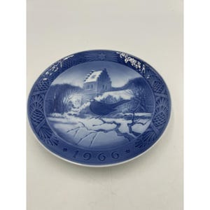 Pode incluir: Prato decorativo azul e branco com uma cena de inverno com um pássaro num galho. O prato tem uma borda detalhada e o ano "1966" é exibido na parte inferior. O prato é redondo e tem um acabamento brilhante.