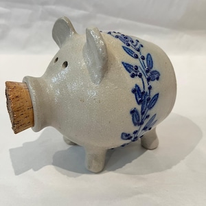 Puede incluir: Una alcancía de cerámica con un tapón de corcho en el hocico. El banco es de color gris claro con diseños florales azules en el lateral. Tiene patas pequeñas y dos agujeros para los ojos.