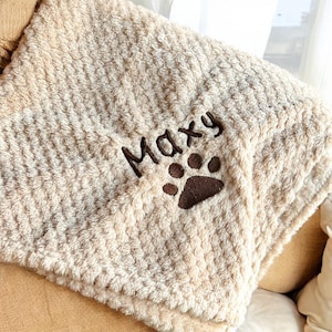 May include: A beige, textured blanket with the name "Maxy" embroidered in dark brown script, accompanied by a matching paw print. The blanket is folded and draped over a light brown surface, suggesting a cozy, pet-friendly item.