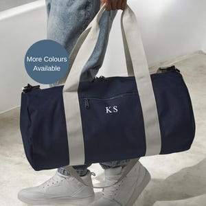 May include: Navy blue duffel bag with light grey straps and the initials "KS" in white. The bag has a zipped front pocket and a cylindrical shape. The text "More Colours Available" is in a blue circle.