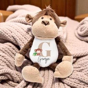 Pode incluir: Um macaco de pelúcia marrom e bege. Ele usa uma camiseta branca com a letra 'G' bege, um desenho de macaco e o nome 'George' em itálico. O brinquedo está em um cobertor rosa. Os braços e as pernas do macaco são marrons.