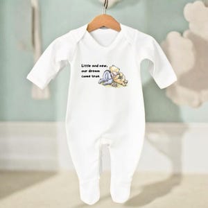 Puede incluir: Mono de bebé blanco con mangas largas, con una ilustración de dibujos animados de Winnie the Pooh y Piglet. El texto "Little and new, our dream come true" está impreso en la parte delantera. El mono está colgado de una percha de madera.