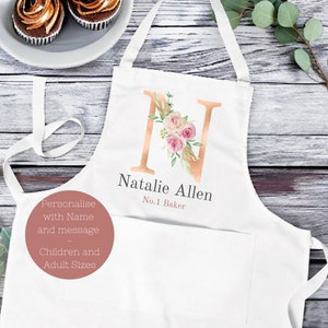 Puede incluir: Delantal blanco con una gran letra "N" color cobre adornada con rosas rosas y vegetación. El delantal tiene el nombre "Natalie Allen" y "No.1 Baker" impreso debajo. Tiene un bolsillo y correas ajustables.
