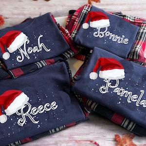 May include: Four navy blue pajama tops with names and Santa hats printed on them. Each top is folded over a red and black plaid pajama bottom. The names are Noah, Bonnie, Pamela, and Reece.