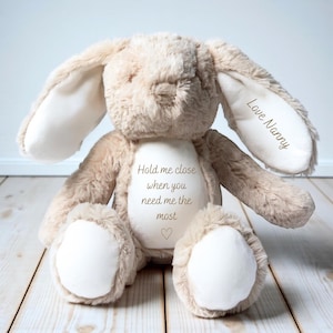 May include: A soft, plush bunny rabbit in beige and cream. The bunny has long, floppy ears and a textured body. Text on the bunny reads "Hold me close when you need me the most" with a heart symbol, and "Love Nanny" on one ear.