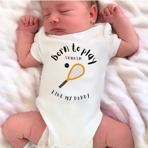 Puede incluir: Mono blanco para bebé con texto negro que dice "Born to Play Squash Like My Daddy" y un gráfico de raqueta de squash negro y naranja.