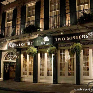 Puede incluir: Una vista nocturna del exterior de un edificio con un letrero que dice "Two Sisters" y "Court of the" sobre la entrada. El edificio tiene un balcón con barandillas de hierro forjado y una puerta negra con una ventana de vidrio. Hay plantas colgando del balcón y del edificio.