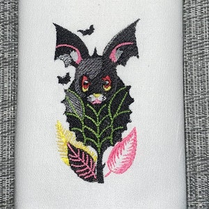 Puede incluir: Toalla de cocina blanca con un murciélago negro bordado con ojos rosas y hojas verdes. El murciélago está volando fuera de un agujero en un árbol.