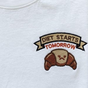 Könnte beinhalten: Ein weißes T-Shirt mit einem gestickten Motiv auf der Brust. Die Stickerei zeigt ein braunes Croissant mit mürrischem Gesichtsausdruck unter einem beigen Banner mit dem schwarzen Text DIET STARTS und dem roten Text TOMORROW.