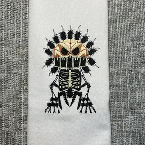 Könnte beinhalten: Weißes Küchentuch mit einem gestickten Design eines Skeletts, das eine Spinnennetzmaske mit Spinnen um den Kopf trägt.