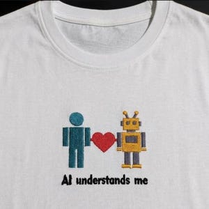 Può includere: T-shirt bianca con un disegno ricamato raffigurante una figura turchese, un robot giallo e un cuore rosso. Il testo "AI understands me" è ricamato sotto. Girocollo.