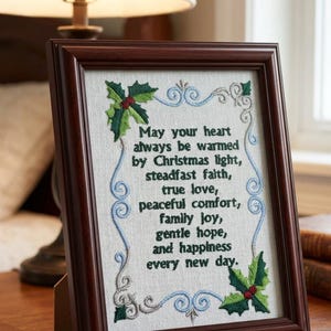 Könnte beinhalten: Ein dunkler Holzrahmen mit einem hellen Stoff, auf dem der Text gestickt ist: May your heart always be warmed by Christmas light, steadfast faith, true love, peaceful comfort, family joy, gentle hope, and happiness every new day.