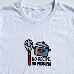 Könnte beinhalten: Weißes T-Shirt mit einem gestickten Design einer Schneebesen und eines Topfes mit einem traurigen Gesicht. Der Text "NO RECIPE, NO PROBLEM" steht darunter. Das Design ist in Schwarz, Rot, Gelb, Blau und Grau gehalten.