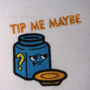 Könnte beinhalten: Gestickter Aufnäher mit einem blauen Glas mit einem mürrischen Gesicht und einem gelben Fragezeichen. Der Text "TIP ME MAYBE" ist oberhalb des Glases in Goldfaden gestickt. Unter dem Glas befindet sich ein goldfarbener Teller.
