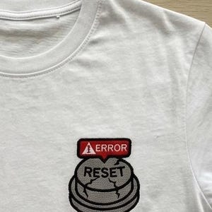 Puede incluir: Una camiseta blanca con un parche bordado en el pecho. El parche muestra un botón circular gris agrietado con la palabra RESET impresa en negro, coronado por un globo de texto rojo con un icono de advertencia blanco y la palabra ERROR.