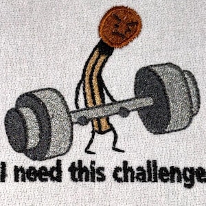 Op de afbeelding: Een geborduurde patch op witte stof met een cartoon luciferfiguurtje met een bruine kop en beige lichaam dat een zware grijze halter optilt. Onder het figuurtje staat de zwarte geborduurde tekst, I need this challenge.