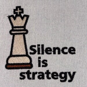 May include: Embroidered chess king piece with the text "Silence is strategy" in black. The chess piece is beige with a black outline and a brown base, set against a light gray fabric background.