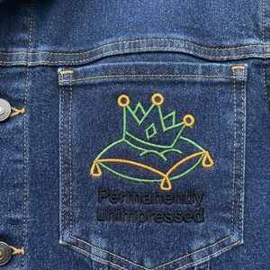 Könnte beinhalten: Eine dunkelblaue Jeansjacke mit einer Brusttasche, die eine Stickerei einer grünen Krone auf einem grün-goldenen Kissen zeigt. Unter der Stickerei ist der Text "Permanently unimpressed" in schwarzem Garn auf die Tasche gestickt.