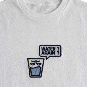 Könnte beinhalten: Weißes T-Shirt mit einem gestickten Design, das ein Wasserglas mit einem Gesicht und einer Sprechblase mit der Aufschrift "WATER? AGAIN?" zeigt. Das Glas ist blau und gelb, der Text ist blau.