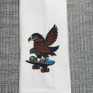 Könnte beinhalten: Weißes Küchentuch mit einem gestickten Design eines schwarzen und roten Adlers, der auf einem blauen Skateboard fährt.