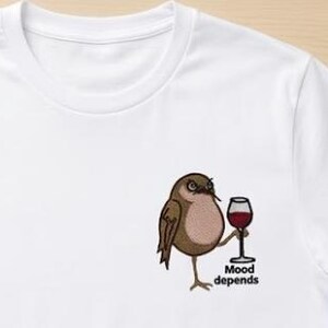 Pode incluir: Camiseta branca com um desenho bordado de um pássaro segurando uma taça de vinho. O pássaro é marrom e bege, e a taça de vinho contém líquido vermelho. O texto "Mood depends" está abaixo do pássaro.