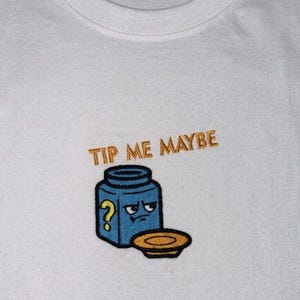 Könnte beinhalten: Weißes T-Shirt mit Rundhalsausschnitt und gesticktem Design. Das Design zeigt ein blaues Glas mit einem mürrischen Gesicht und einem gelben Fragezeichen sowie den Text "TIP ME MAYBE" in Goldfaden.