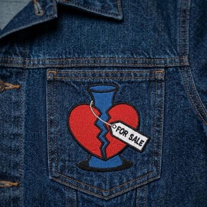 May include: A dark blue denim jacket featuring an embroidered patch on the chest pocket. The patch depicts a red broken heart shaped like a vase with a blue base and neck, accompanied by a white tag with the black text FOR SALE attached.
