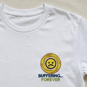 Könnte beinhalten: Ein weißes T-Shirt mit einem gestickten Motiv auf der Brust. Das Design besteht aus einem gelben, kreisförmigen Ladesymbol mit einem traurigen Gesicht in der Mitte, ergänzt durch den Text "BUFFERING..." in Dunkelblau und "FOREVER" in Grün.
