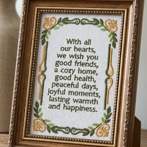 Könnte beinhalten: Ein gerahmtes Stickbild mit dem Text: With all our hearts, we wish you good friends, a cozy home, good health, peaceful days, joyful moments, lasting warmth and happiness. Das Design enthält grüne und goldene florale Bordüren.