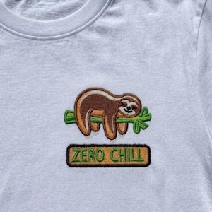 Könnte beinhalten: Weißes T-Shirt mit einem braunen und beigen Faultier, das auf einem grünen Ast gestickt ist. Darunter befindet sich ein rechteckiger Aufnäher mit dem grünen Schriftzug "ZERO CHILL".