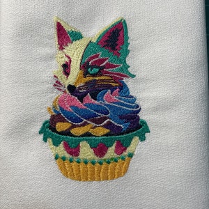Könnte beinhalten: Ein farbenfrohes besticktes Geschirrtuch mit einem Fuchs, der in einem Cupcake sitzt. Der Fuchs ist aus rosa, grünem, gelbem und blauem Garn gefertigt. Der Cupcake ist gelb, grün und rosa.