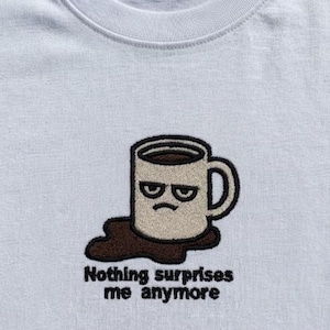 Puede incluir: Camiseta blanca con un diseño bordado de una taza de café gruñona con café derramado y el texto "Nothing surprises me anymore" en negro. La taza tiene un contorno marrón oscuro y está llena de café.