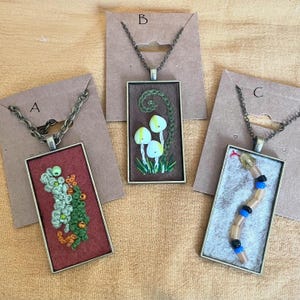 May include: Three necklaces with different pendants. The first pendant is a red rectangle with green and orange embroidery. The second pendant is a brown rectangle with green and white embroidery of mushrooms. The third pendant is a white rectangle with a brown snake made of beads.