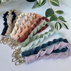 Puede incluir: Una colección de nueve llaveros de pulsera de macramé en varios colores, incluyendo negro, beige, crema, rosa empolvado, terracota, verde salvia, azul claro, azul marino y lavanda. Cada llavero presenta un diseño de cordón trenzado y un cierre de metal plateado.