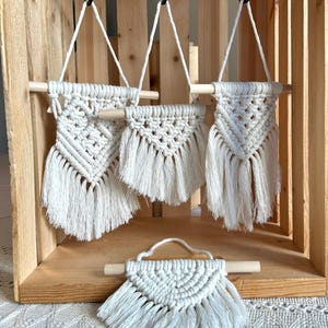 Könnte beinhalten: Eine Sammlung cremefarbener Makramee-Wandbehänge mit geometrischem Muster und Fransen. Die Wandbehänge sind an Holzstäben und schwarzen Haken aufgehängt und in einer Holzkiste ausgestellt. Das kleinste Makramee-Stück liegt auf einer Oberfläche.